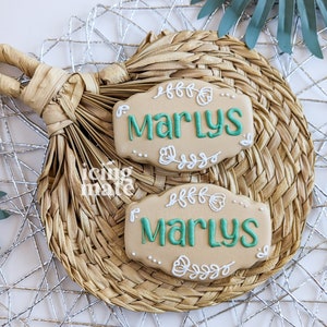 Pu&ograve; includere: Due biscotti beige con il nome "Marlys" in glassa verde acqua, circondati da disegni floreali bianchi. I biscotti poggiano su un sottobicchiere beige intrecciato con un manico, su uno sfondo di filo d'argento e una foglia verde.