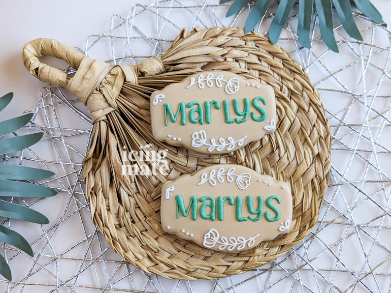 Pu&ograve; includere: Due biscotti beige con il nome "Marlys" in glassa verde acqua, circondati da disegni floreali bianchi. I biscotti poggiano su un sottobicchiere beige intrecciato con un manico, su uno sfondo di filo d'argento e una foglia verde.
