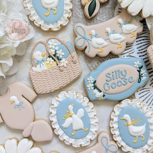 May include: Assortment of decorated sugar cookies with a baby shower theme. Cookies include a onesie, daisies, and oval cookies with a goose design. Colors include white, light blue, yellow, and tan.