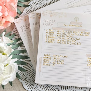 May include: A white order form with a gold floral design and the words "Order Form" and "Fox Woodland" printed on it. The form lists different cookie designs and colors with corresponding quantities and prices.