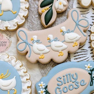 May include: Assortment of decorated sugar cookies with a whimsical goose theme. Cookies feature white geese with blue bows, set against light blue and beige backgrounds. Some cookies have the words "Silly Goose".
