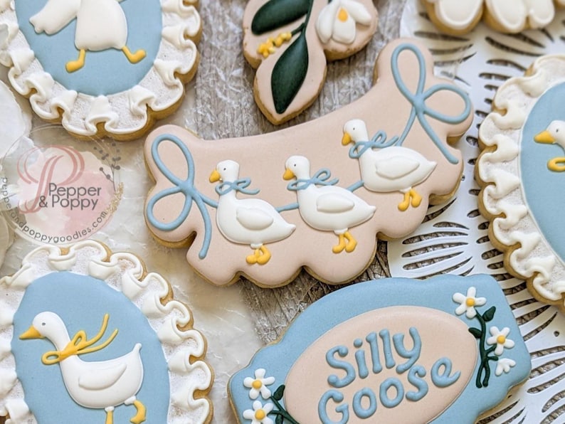 May include: Assortment of decorated sugar cookies with a whimsical goose theme. Cookies feature white geese with blue bows, set against light blue and beige backgrounds. Some cookies have the words "Silly Goose".