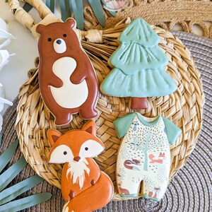 Pu&ograve; includere: Quattro biscotti di zucchero decorati disposti su un vassoio intrecciato. I biscotti hanno la forma di un orso, un albero di Natale, una volpe e un body per neonati. L'orso &egrave; marrone con la pancia bianca. L'albero &egrave; verde. La volpe &egrave; arancione e bianca. Il body ha un design boschivo.