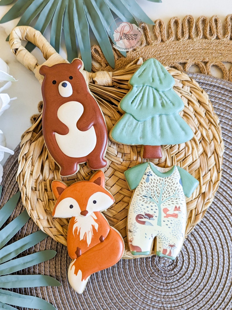 Pu&ograve; includere: Quattro biscotti di zucchero decorati disposti su un vassoio intrecciato. I biscotti hanno la forma di un orso, un albero di Natale, una volpe e un body per neonati. L'orso &egrave; marrone con la pancia bianca. L'albero &egrave; verde. La volpe &egrave; arancione e bianca. Il body ha un design boschivo.