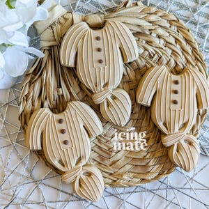 May include: Four beige-colored baby onesie cookies with vertical stripes and brown button details, arranged on a woven basket. The cookies are decorated with icing and have a bow-tie design at the bottom. White flowers are in the background.