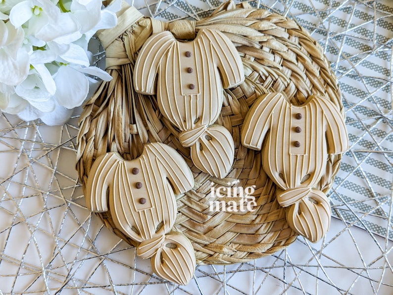 May include: Four beige-colored baby onesie cookies with vertical stripes and brown button details, arranged on a woven basket. The cookies are decorated with icing and have a bow-tie design at the bottom. White flowers are in the background.