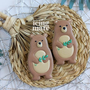 Pu&ograve; includere: Due biscotti a forma di orso con glassa marrone, pancia beige e occhi neri. Ogni orso tiene in mano un rametto di foglie verdi. I biscotti sono su un sottobicchiere circolare intrecciato con uno sfondo geometrico argentato. Il testo "icing mate" &egrave; visibile.