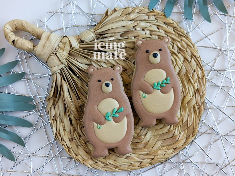 Pu&ograve; includere: Due biscotti a forma di orso con glassa marrone, pancia beige e occhi neri. Ogni orso tiene in mano un rametto di foglie verdi. I biscotti sono su un sottobicchiere circolare intrecciato con uno sfondo geometrico argentato. Il testo "icing mate" &egrave; visibile.