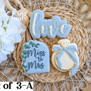 May include: Set of three decorated cookies. One cookie says "love" in white script on a blue cloud shape. Another says "Miss to Mrs" with floral accents. The third is a ring with a diamond and blue bow.