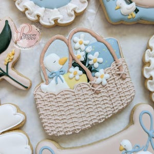May include: Decorated sugar cookies with a baby shower theme. The cookies feature a basket with a goose, daisies, and eggs, along with other cookies with geese and floral designs. The cookies are decorated with pastel colors and intricate details.