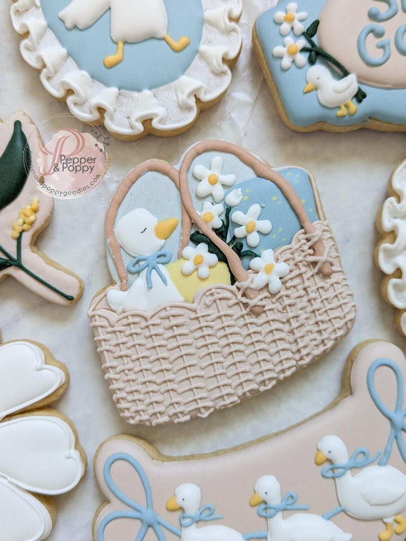 May include: Decorated sugar cookies with a baby shower theme. The cookies feature a basket with a goose, daisies, and eggs, along with other cookies with geese and floral designs. The cookies are decorated with pastel colors and intricate details.