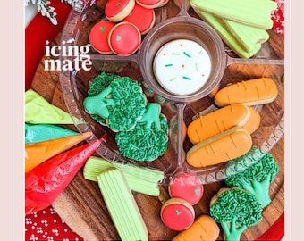 May include: A clear plastic divided tray filled with decorated sugar cookies shaped like vegetables. Cookies include tomatoes, carrots, broccoli, and celery. The tray also contains a round cookie with sprinkles. The text "icing mate" is visible.