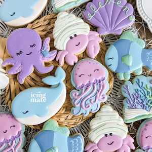 May include: A collection of decorated sugar cookies featuring various sea creatures, including an octopus, a whale, a crab, a jellyfish, and a fish. The cookies are decorated with icing and have a light blue, purple, and green color scheme. The text "icing mate" is written on one of the cookies.