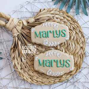 May include: Two beige cookies with the name "Marlys" in teal script, decorated with white floral designs. The cookies rest on a woven straw placemat, with a silver wire mesh and a green leaf in the background.