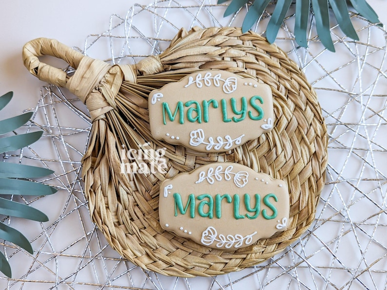 May include: Two beige cookies with the name "Marlys" in teal script, decorated with white floral designs. The cookies rest on a woven straw placemat, with a silver wire mesh and a green leaf in the background.