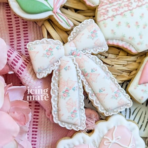 May include: Close-up of decorated sugar cookies. The cookies are in the shape of a bow, a baby outfit, a heart, and a floral bouquet. The bow cookie has a floral pattern with pink bows. The text "icing mate" is visible.