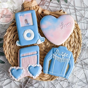 May include: Four decorated sugar cookies on a woven placemat. The cookies are decorated with blue and pink icing and feature a music note, a heart with the word "Lover", a blue denim jacket with the word "Swift", and a blue iPod with the word "Lover" and a picture of a woman.