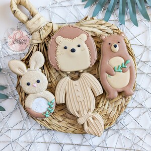 Pu&ograve; includere: Una collezione di biscotti decorati a forma di coniglio, riccio, orso e tutina per neonati. I biscotti sono marrone chiaro con dettagli bianchi e pastello, disposti su un sottobicchiere intrecciato, creando un'estetica naturale e rustica.
