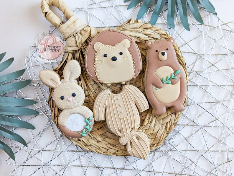Pu&ograve; includere: Una collezione di biscotti decorati a forma di coniglio, riccio, orso e tutina per neonati. I biscotti sono marrone chiaro con dettagli bianchi e pastello, disposti su un sottobicchiere intrecciato, creando un'estetica naturale e rustica.