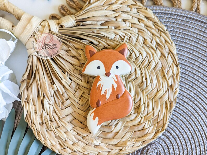 Pu&ograve; includere: Un biscotto a forma di volpe decorato con glassa arancione, bianca e marrone. Il biscotto &egrave; posto su un sottobicchiere intrecciato di colore naturale. Il biscotto ha un design dettagliato, con pancia e punta della coda bianche. Una dolcezza.