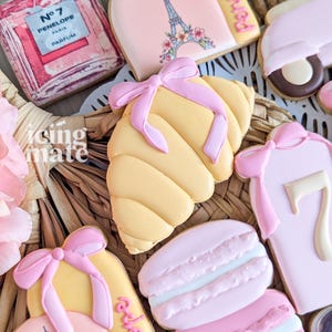 May include: Assortment of decorated sugar cookies in a basket. Cookies include a croissant with a pink bow, a perfume bottle, Eiffel Tower, and macarons. The cookies are primarily pink, yellow, and white.