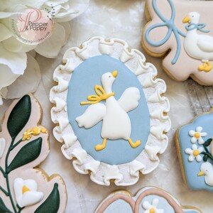 May include: Assortment of decorated sugar cookies with a baby shower theme. One cookie features a white goose with a yellow bow on a light blue oval, framed by white frosting. Other cookies have floral and bow designs.