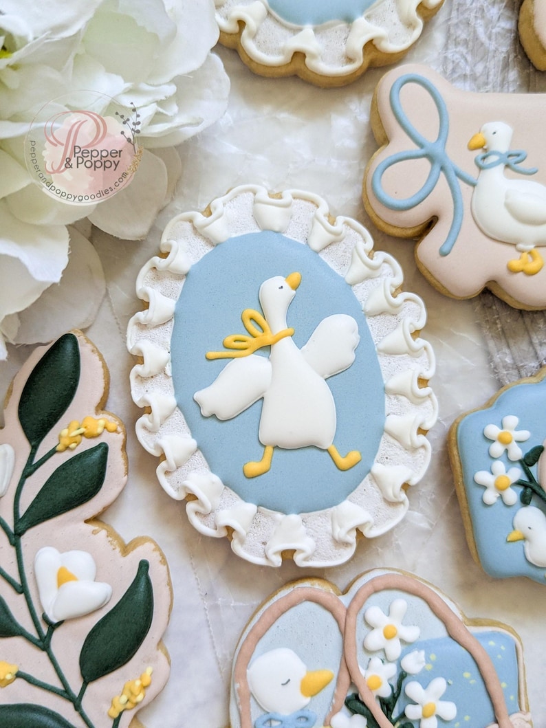 May include: Assortment of decorated sugar cookies with a baby shower theme. One cookie features a white goose with a yellow bow on a light blue oval, framed by white frosting. Other cookies have floral and bow designs.