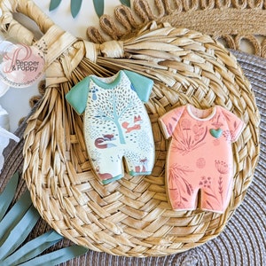 Pu&ograve; includere: Due biscotti a forma di tutina per neonati. Uno &egrave; verde chiaro e bianco con una scena della foresta e volpi. L'altro &egrave; color pesca con un disegno floreale e un cuore verde. Entrambi i biscotti sono su un tappetino intrecciato.
