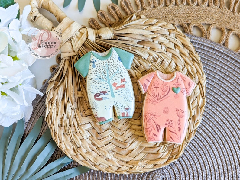 Pu&ograve; includere: Due biscotti a forma di tutina per neonati. Uno &egrave; verde chiaro e bianco con una scena della foresta e volpi. L'altro &egrave; color pesca con un disegno floreale e un cuore verde. Entrambi i biscotti sono su un tappetino intrecciato.