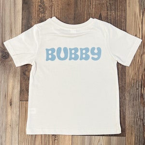 May include: A white t-shirt with a blue retro-style font that says "BUBBY" on the back.