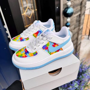 May include: White sneakers with a blue trim and a colorful puzzle piece pattern on the upper. The sneakers are in a white box.
