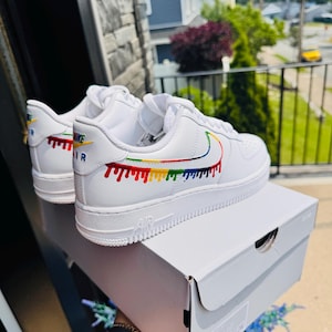 May include: White Nike Air Force 1 sneakers with a rainbow drip design on the side and heel. The Nike swoosh is outlined in rainbow colors. The shoes are displayed on top of a white shoe box.