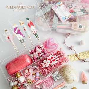 Puede incluir: Cajas organizadoras de plástico transparente llenas de artículos decorativos rosas, blancos y dorados. Incluye plastilina rosa, cuentas, bastones de caramelo, árboles de Navidad en miniatura y recortes de papel con temática de Cascanueces. Las cajas están atadas con cinta plateada.