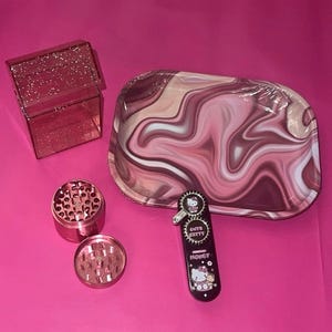 May include: A pink glittery cigarette case, a rectangular rolling tray with a pink and white marble swirl pattern, a two-piece pink metal herb grinder, and a black lighter featuring a Hello Kitty graphic and the text Cute Kitty Honey.