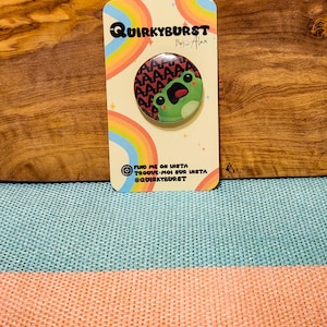 Peut inclure: Épingle ronde avec un motif de grenouille verte et le mot "AAAAAAA" dans un motif rouge. L'épingle est sur une carte avec le texte "QUIRKYBURST By Alex" et des réseaux sociaux. Un arc-en-ciel est en arrière-plan.