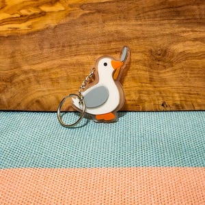 May include: A whimsical keychain featuring a cartoon seagull design. The seagull has a white body, gray wings, and orange beak and feet. The keychain is attached to a silver metal ring and chain. The background is a wooden surface and a blue and peach fabric.