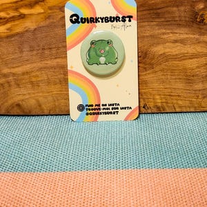 Peut inclure: Un badge rond représentant une grenouille verte souriante sur fond vert clair. L'épingle est fixée à une carte crème avec un motif arc-en-ciel et le texte "Quirkyburst". La carte comprend également des identifiants de réseaux sociaux.