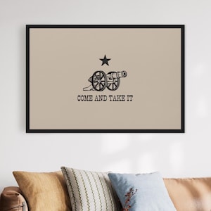 Puede incluir: Una impresión enmarcada de la bandera "Come and Take It", que presenta un cañón negro y una sola estrella blanca sobre un fondo beige. El texto "Come and Take It" está impreso en negro debajo del cañón.