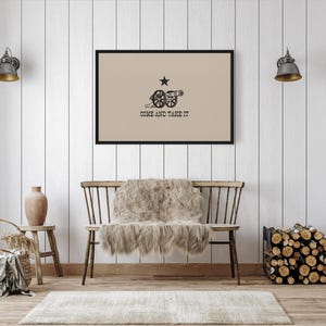 May include: A framed print of the "Come and Take It" flag, featuring a cannon and a lone star on a beige background. The flag is displayed on a white wood-paneled wall above a wooden bench with a faux fur throw.