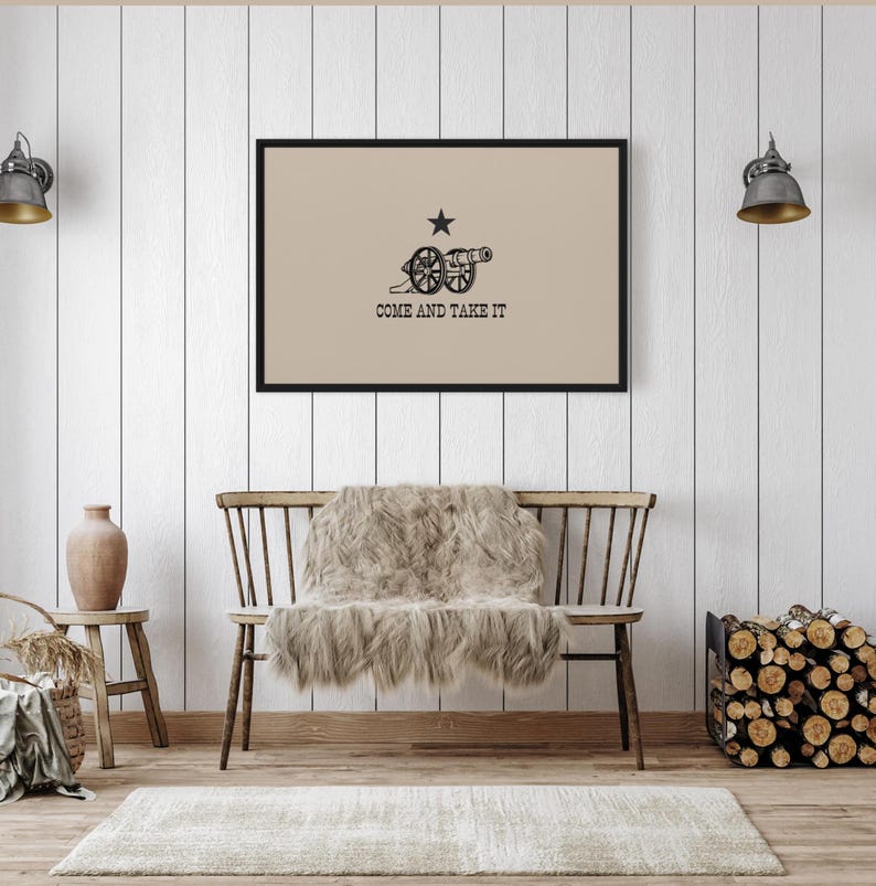 May include: A framed print of the "Come and Take It" flag, featuring a cannon and a lone star on a beige background. The flag is displayed on a white wood-paneled wall above a wooden bench with a faux fur throw.