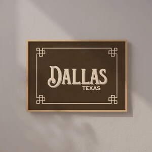 Pu&ograve; includere: Un cartello incorniciato con la parola "DALLAS" in un font vintage e "TEXAS" sotto. Il cartello ha uno sfondo marrone scuro, un bordo chiaro e una cornice in legno. Elementi decorativi angolari completano il design.