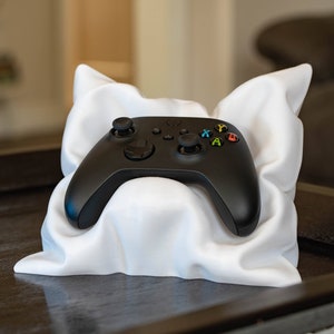 May include: A black Xbox controller resting on a white pillow. The controller has a black and green color scheme with buttons labeled A, B, X, and Y.