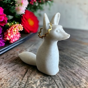 May include: A white ceramic fox figurine with a gold zig-zag ring around its neck. The fox is sitting on a wooden surface.