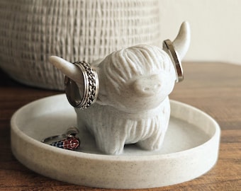 May include: White ceramic ring holder shaped like a Highland cow with a speckled finish. The cow is holding three silver rings on its horns.