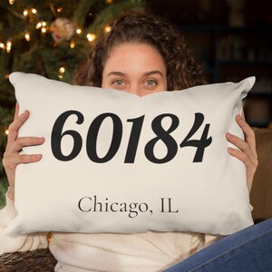Peut inclure: Coussin rectangulaire crème avec les chiffres noirs "60184" et le texte "Chicago, IL". Le coussin est tenu devant une personne, avec un sapin de Noël en arrière-plan.