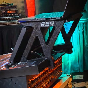 May include: A black, textured laptop stand with the letters RSR printed in white on the side. The stand is elevated and holds a laptop above a piece of audio equipment with numerous circular ports and orange glowing lights in a dark setting.