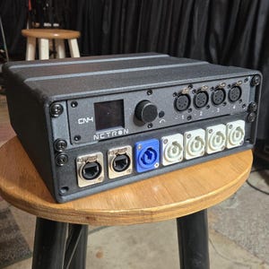 Puede incluir: Un dispositivo electrónico negro con múltiples puertos de entrada y salida, una pequeña pantalla y un mando giratorio. El dispositivo tiene el texto "CN4" y "NETRON" impreso. Está sobre un taburete de madera.