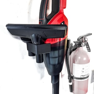 Pode incluir: Um aspirador de pó de mão preto e vermelho está montado em um suporte de parede. O aspirador de pó tem uma haste de extensão longa e preta e um bico preto. O suporte é feito de plástico preto e tem duas ranhuras para os acessórios do aspirador de pó. Um extintor de incêndio vermelho está montado na parede ao lado do aspirador de pó.