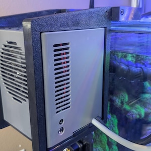 Peut inclure: Un refroidisseur d'aquarium gris et noir avec un ventilateur et une bouche d'aération sur le côté. Le refroidisseur est monté sur le côté d'un aquarium avec une lumière bleue.