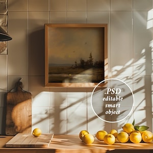 Può includere: Un dipinto di paesaggio incorniciato con una cornice in legno è appeso a una parete piastrellata di bianco sopra un bancone da cucina. Il bancone è in legno e ha un tagliere in legno e una ciotola di limoni. Il dipinto è un paesaggio con un fiume e alberi. Il testo "PSD editable smart object" è scritto in un cerchio sull'immagine.